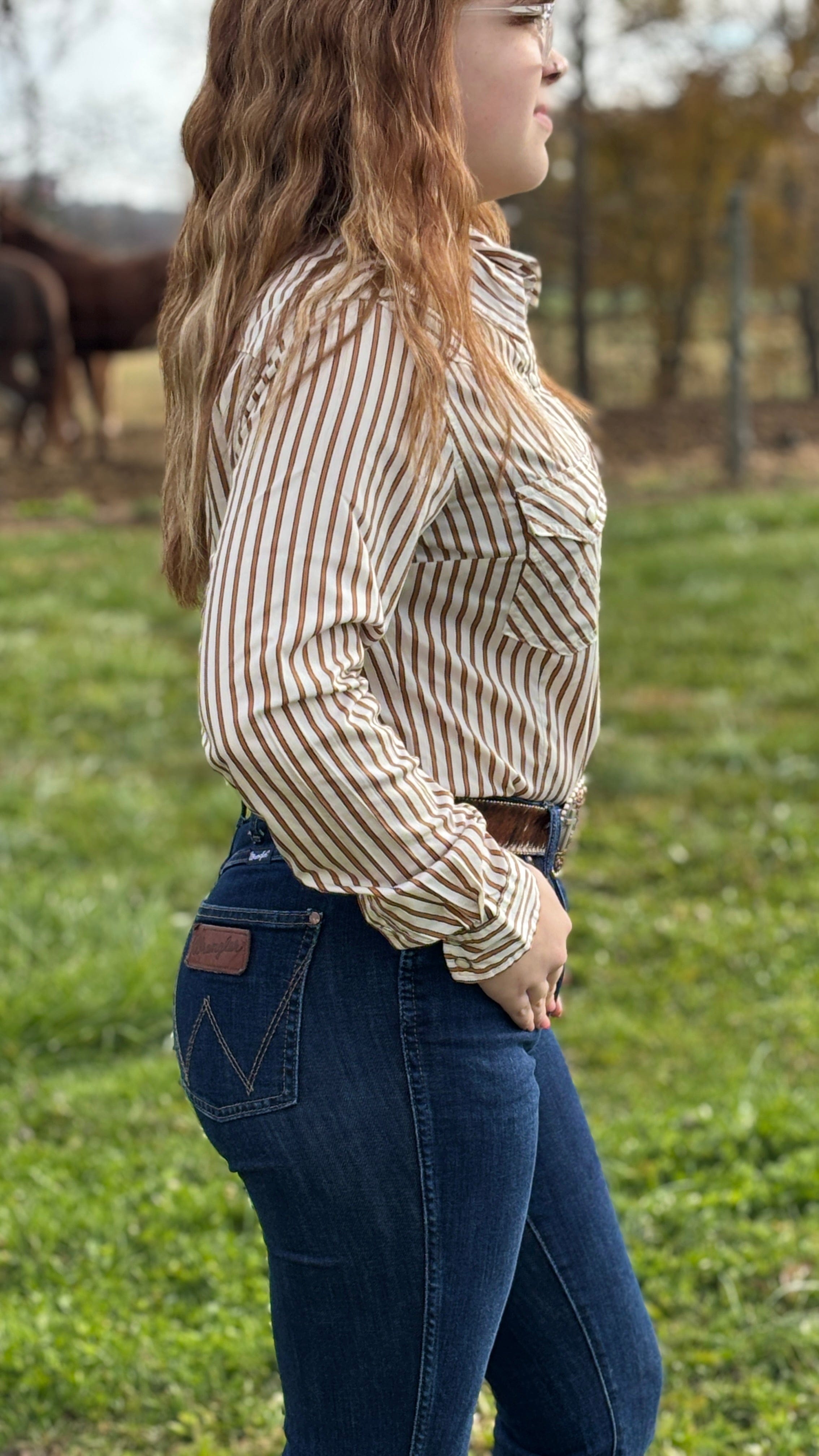 Woman wearing a striped shirt and jeans standing in a grassy field with horses in the background